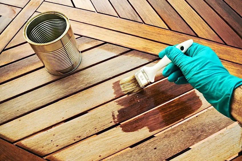 A person wearing gloves cleans a wooden deck with a brush, applying the sealer