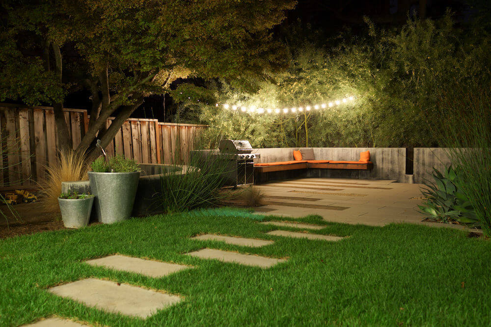 A backyard with a patio, grill, and outdoor lighting
