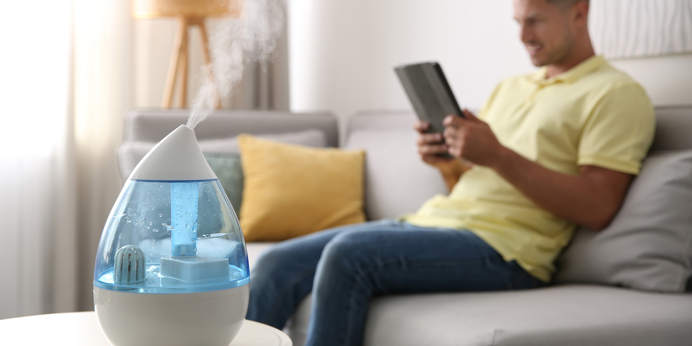 A man sitting on a couch, using a tablet, with a humidifier nearby. Steam Inhalers.